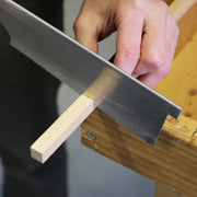 Kakuri Dozuki Japanese hand Saw with a wood handle wrapped in rattan and metal saw blade with fine teeth being used to cut a square wooden dowel on a wooden workbench